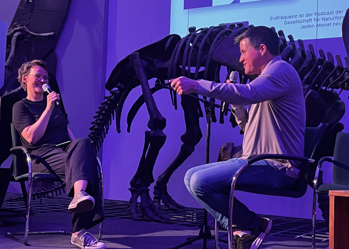 Zwei Personen auf eine Bühne im Gespräch. Im Hintergrund das Skellett eines Dinosauriers.
